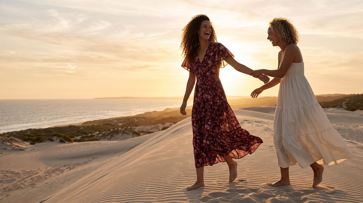 Two friends walking a backlit dune ridge at sunset in a burgundy floral maxi dress and an ivory tiered maxi dress