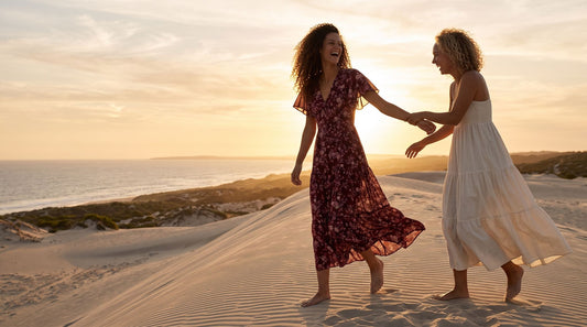 Two friends walking a backlit dune ridge at sunset in a burgundy floral maxi dress and an ivory tiered maxi dress